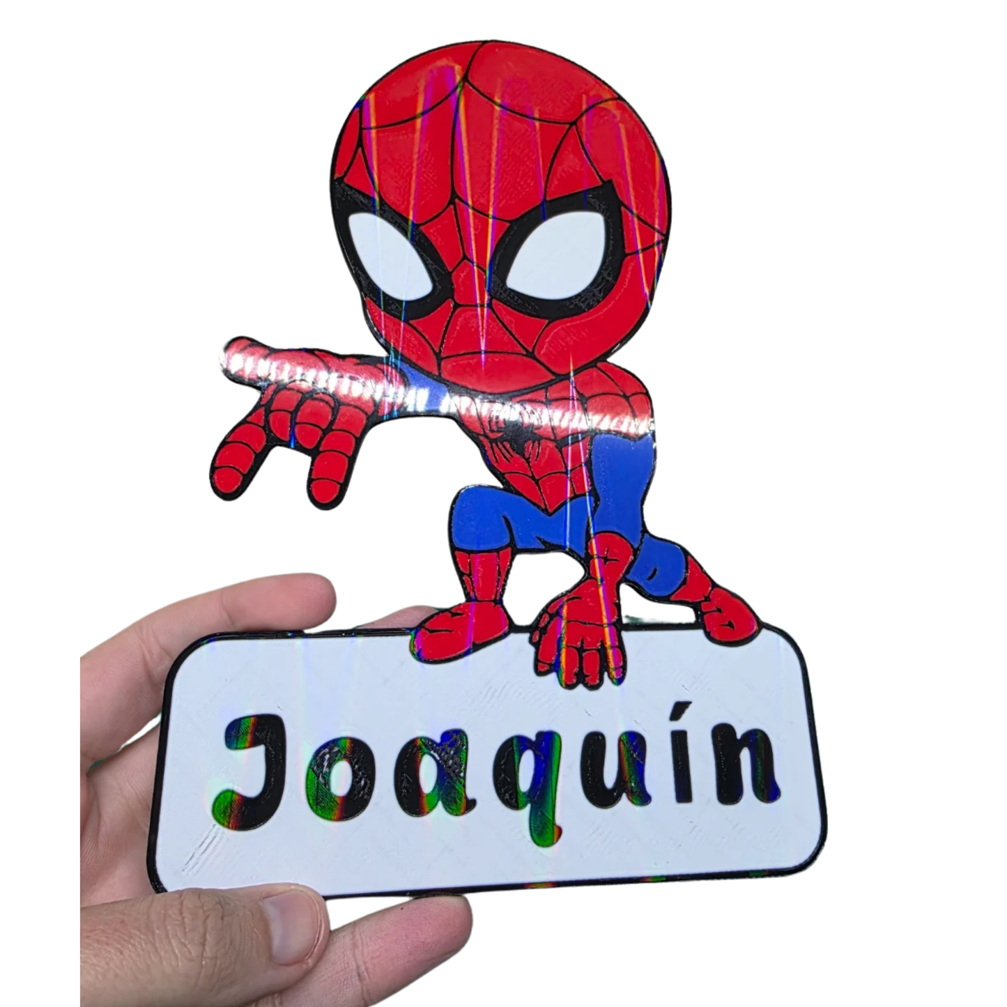Mano sosteniendo un aplique decorativo de Spider-Man chibi con el nombre "personalizado" en la base, mostrando su tamaño y detalle.