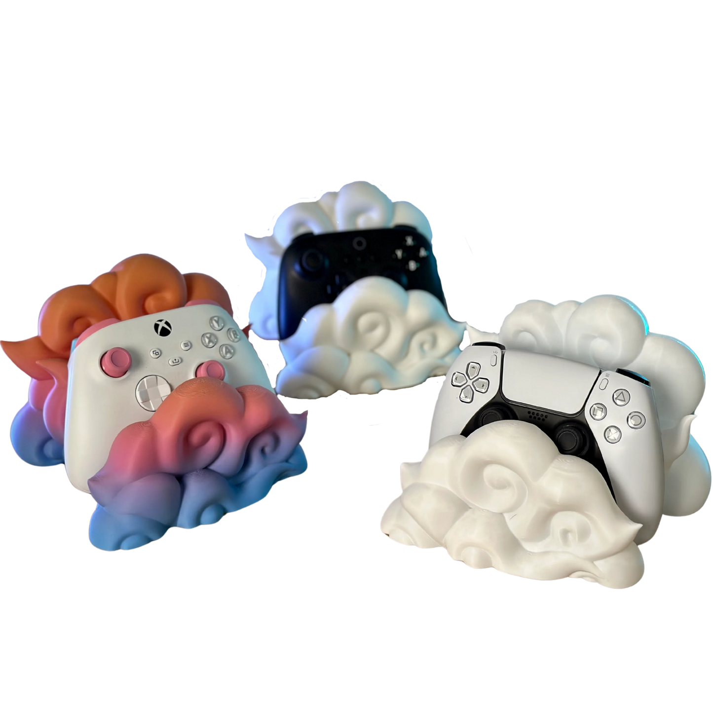 Colección de tres soportes para mandos gaming en forma de nube (blanco, negro, degradado rosa/azul) con mandos PS5 y Xbox, mostrando variedad de estilos.