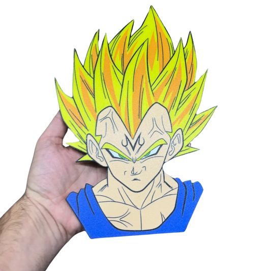 Aplique decorativo de la cabeza de Majin Vegeta de Dragon Ball Z, con cabello Super Saiyajin amarillo y la "M" en la frente, siendo sostenido por una mano.
