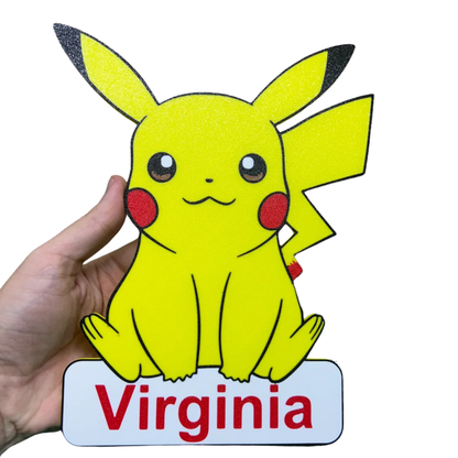 Aplique decorativo de Pikachu Pokémon en color amarillo, con mejillas rojas y cola de rayo, sobre una base con el nombre "personalizado", sostenido por una mano.