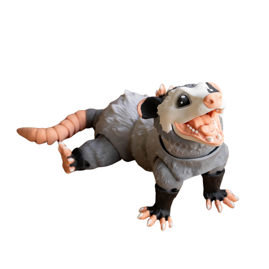 Figura articulada de zarigüeya en color gris, con boca abierta, posada en el suelo simulando la pose de "hacerse el muerto" (playing possum)