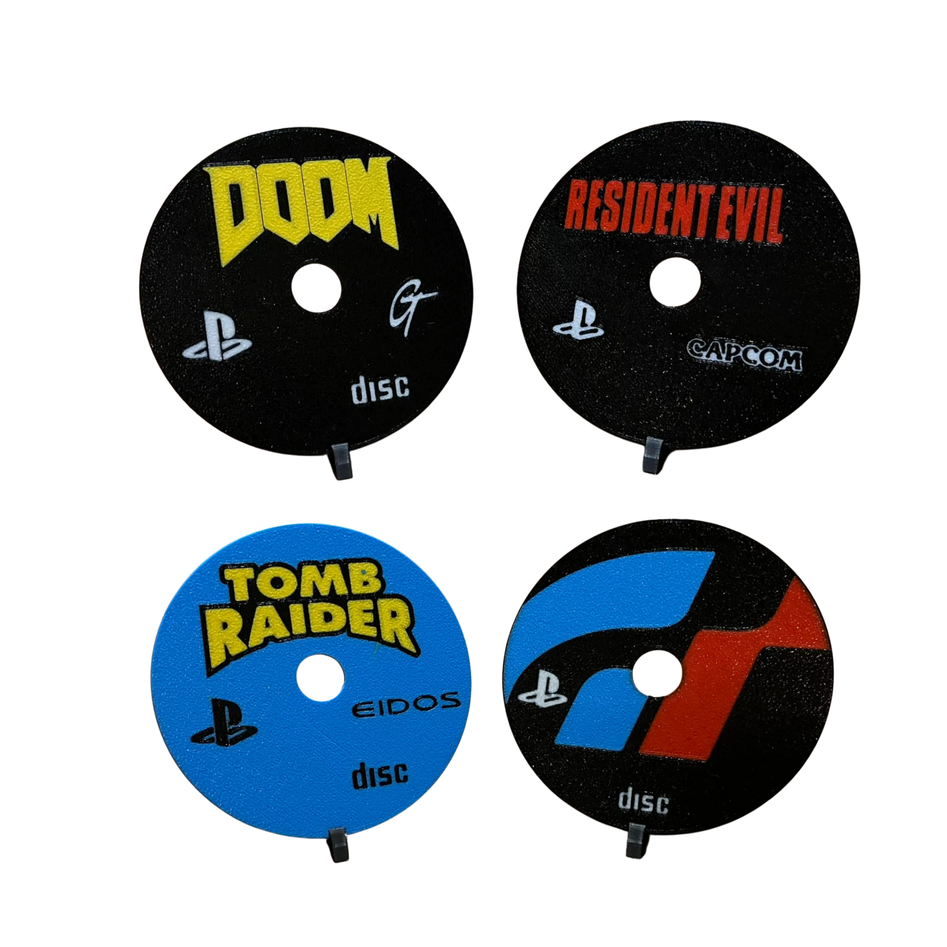Cuatro posavasos de discos de PlayStation 1: DOOM (amarillo y negro), Resident Evil (negro), Tomb Raider (azul) y Gran Turismo (azul y rojo).