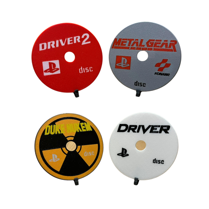 Cuatro posavasos de discos de PlayStation 1: Driver 2 (rojo), Metal Gear Solid (gris), Duke Nukem (amarillo y negro) y Driver (blanco).