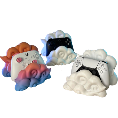 Colección de tres soportes para mandos gaming en forma de nube (blanco, negro, degradado rosa/azul) con mandos PS5 y Xbox, mostrando variedad de estilos.